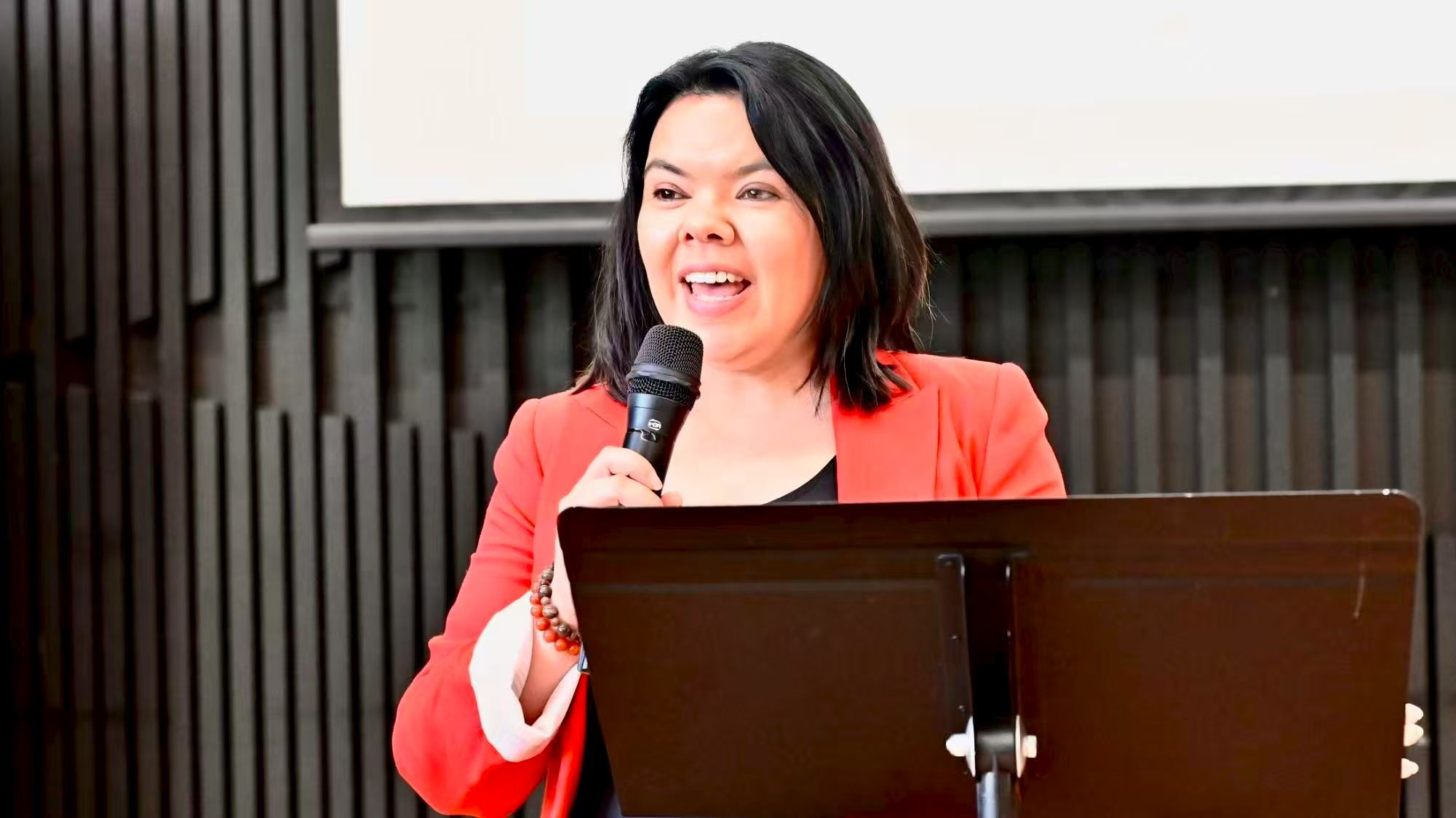 A speaker wearing a red jacket addressing the gathering at the lectern