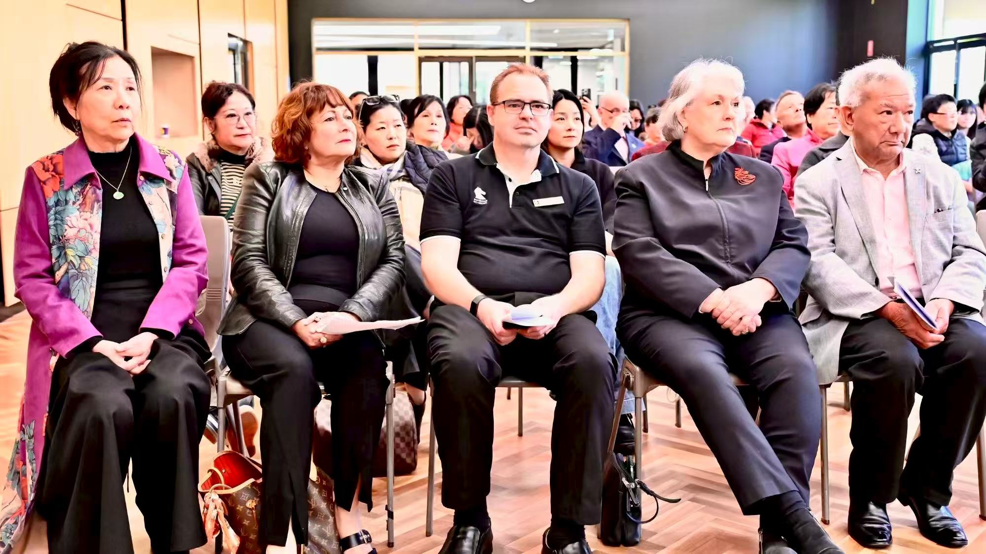 Front-row guests holding event programmes and listening to a speaker